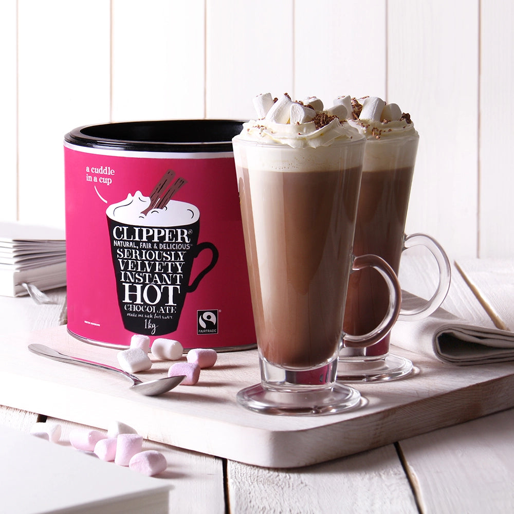 Bulk tub of hot chocolate powder from Clipper next to two tall latte glasses of hot chocolate topped with whipped cream and marshmallows